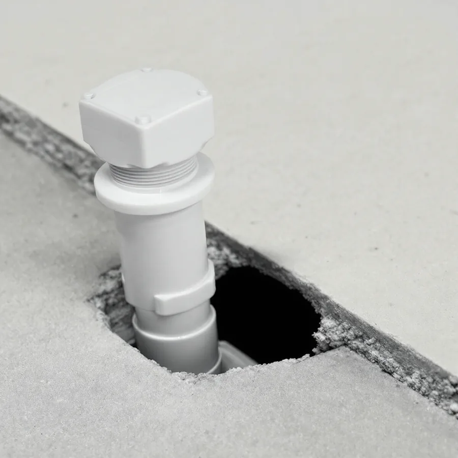White plastic drain pipe with a round white threaded cap with a raised square nub rising from a concrete basement floor