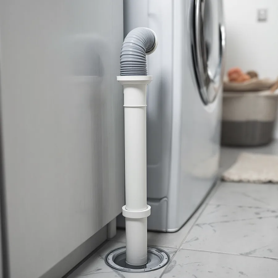 White plastic vertical standpipe rising from the floor behind a washing machine with a grey corrugated drain hose hooked over its top and a small puddle at its base