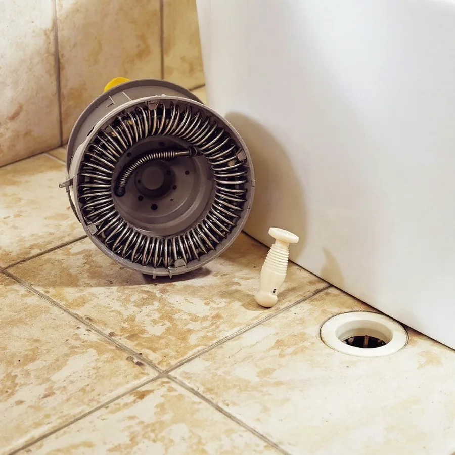 Hand crank drain auger with a coiled steel cable in a round drum lying on a tiled bathroom floor beside a bathtub drain