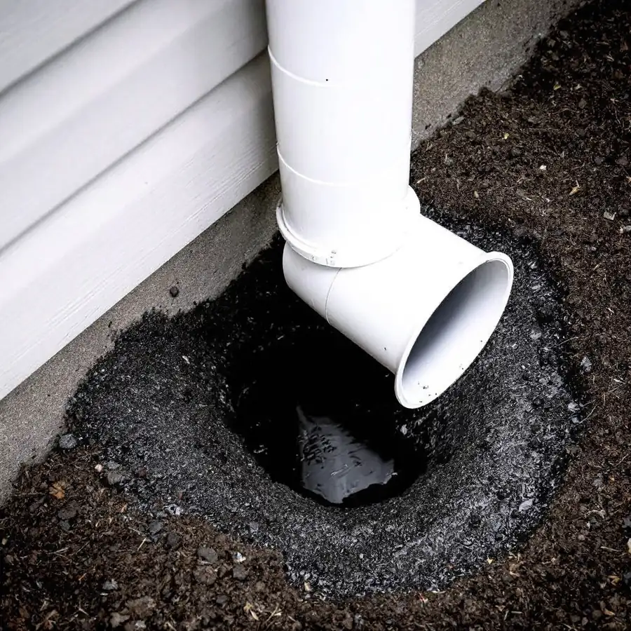 The bottom of a white downspout ending a few inches above bare soil right against a house foundation, with a washed-out hollow and standing water in the ground beneath it