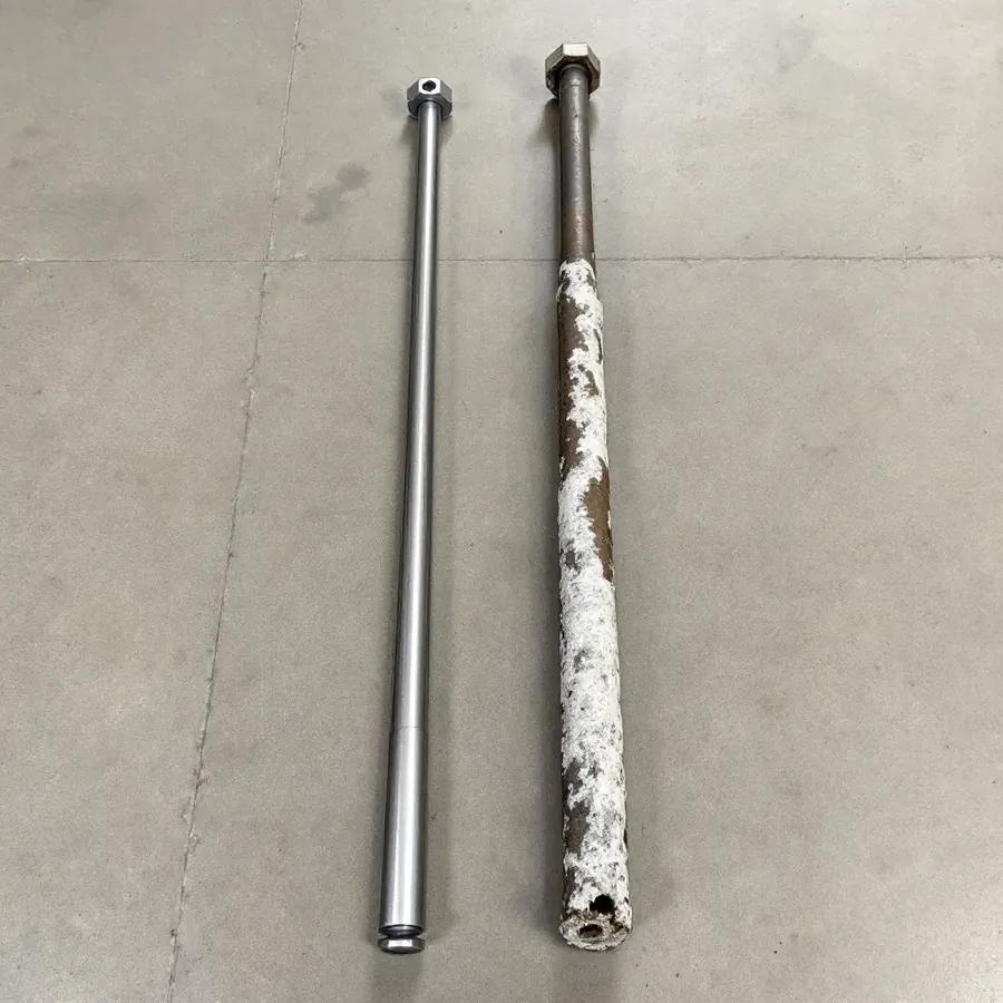Two long thin metal rods side by side on a concrete floor, one new and smooth with a hexagonal head, the other eaten to a thin pitted core with white crust